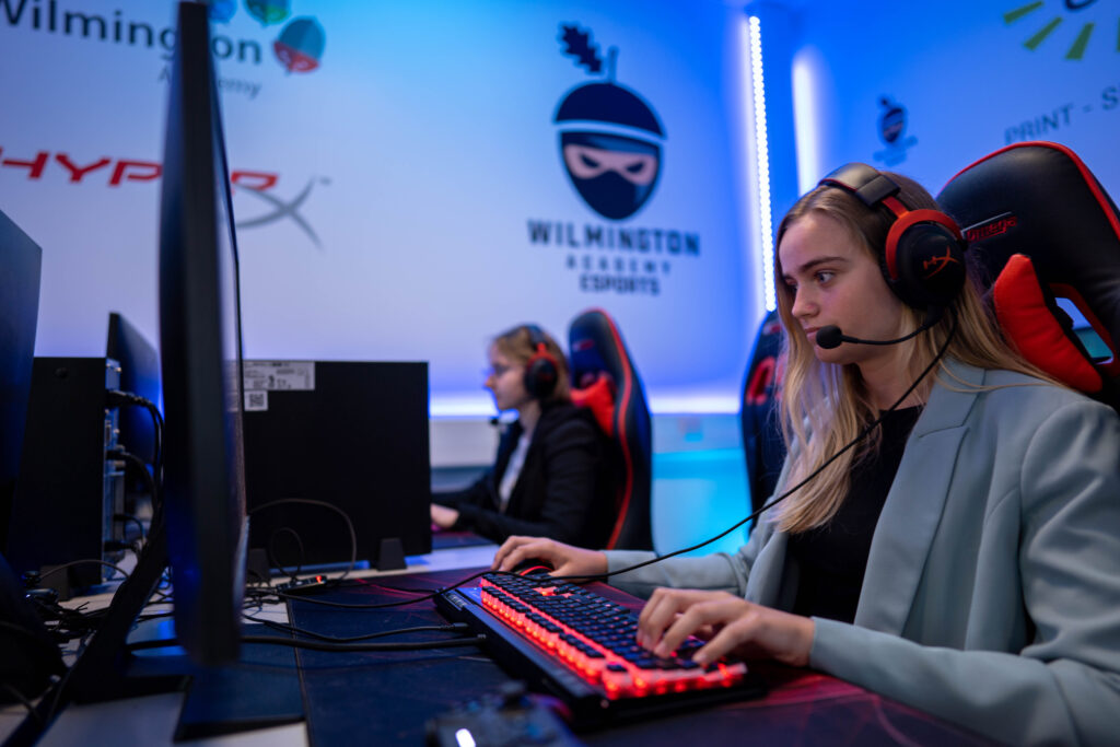 Two students wearing headphones, working at computers, in front of a wall with the Wilmington eSports logo on it