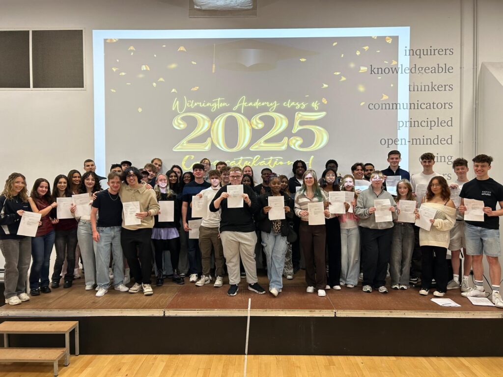 A shot of students on the stage, all holding their certificates and smiling. The "Wilmington Academy class of 2025 Congratulations!" message is prominent on the screen behind them. On the right side of the wall, there are words like "inquirers," "knowledgeable," "thinkers," "communicators," "principled," and "open-minded," which might be related to the academy's values.