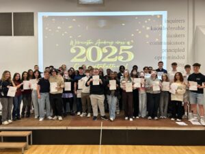 A shot of students on the stage, all holding their certificates and smiling. The "Wilmington Academy class of 2025 Congratulations!" message is prominent on the screen behind them. On the right side of the wall, there are words like "inquirers," "knowledgeable," "thinkers," "communicators," "principled," and "open-minded," which might be related to the academy's values.
